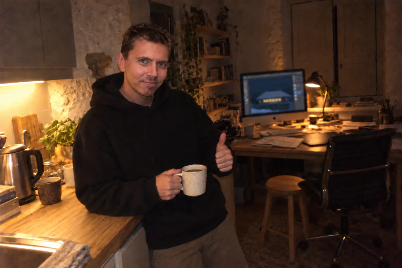 Hal in a black hoodie leaning on the kitchen worktop in the barn studio late at night, a mug of coffee in one hand and a thumbs up with the other, a snowy night scene open in Illustrator on the iMac behind him.